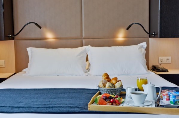 A neatly made bed with white linens, a tray with breakfast including croissants, fruit, and coffee, and a bedside table with a phone and a lamp.
