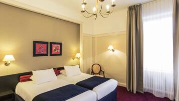 A hotel room with two beds, a chandelier, wall-mounted lamps, and a window with curtains.