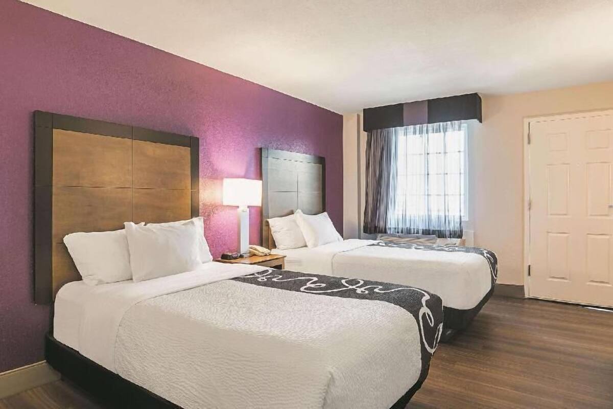 A hotel room with two beds, a large headboard, a bedside table with a lamp, and a window with curtains.