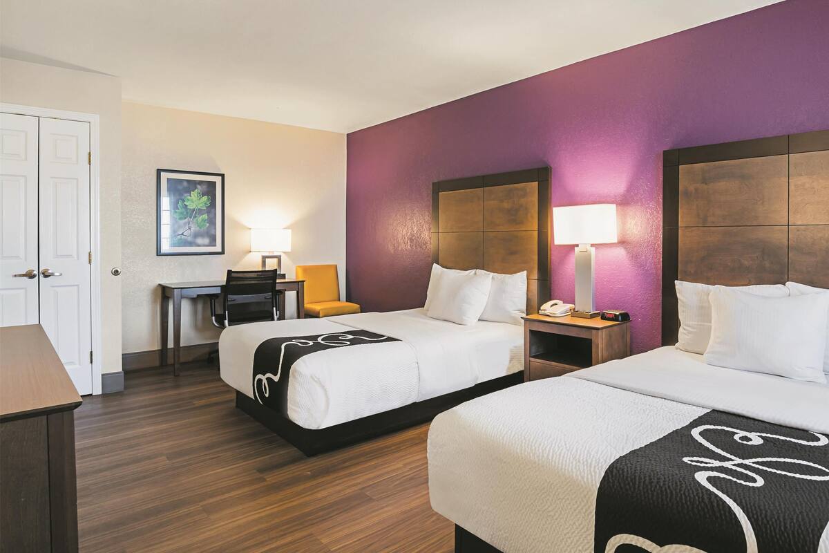 A hotel room with two beds, a desk, a chair, and a nightstand with a lamp.