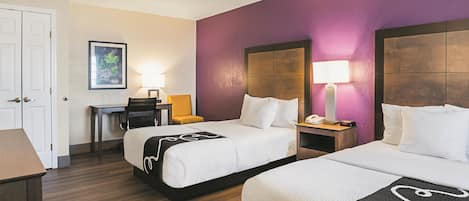 A hotel room with two beds, a desk, a chair, and a nightstand with a lamp.