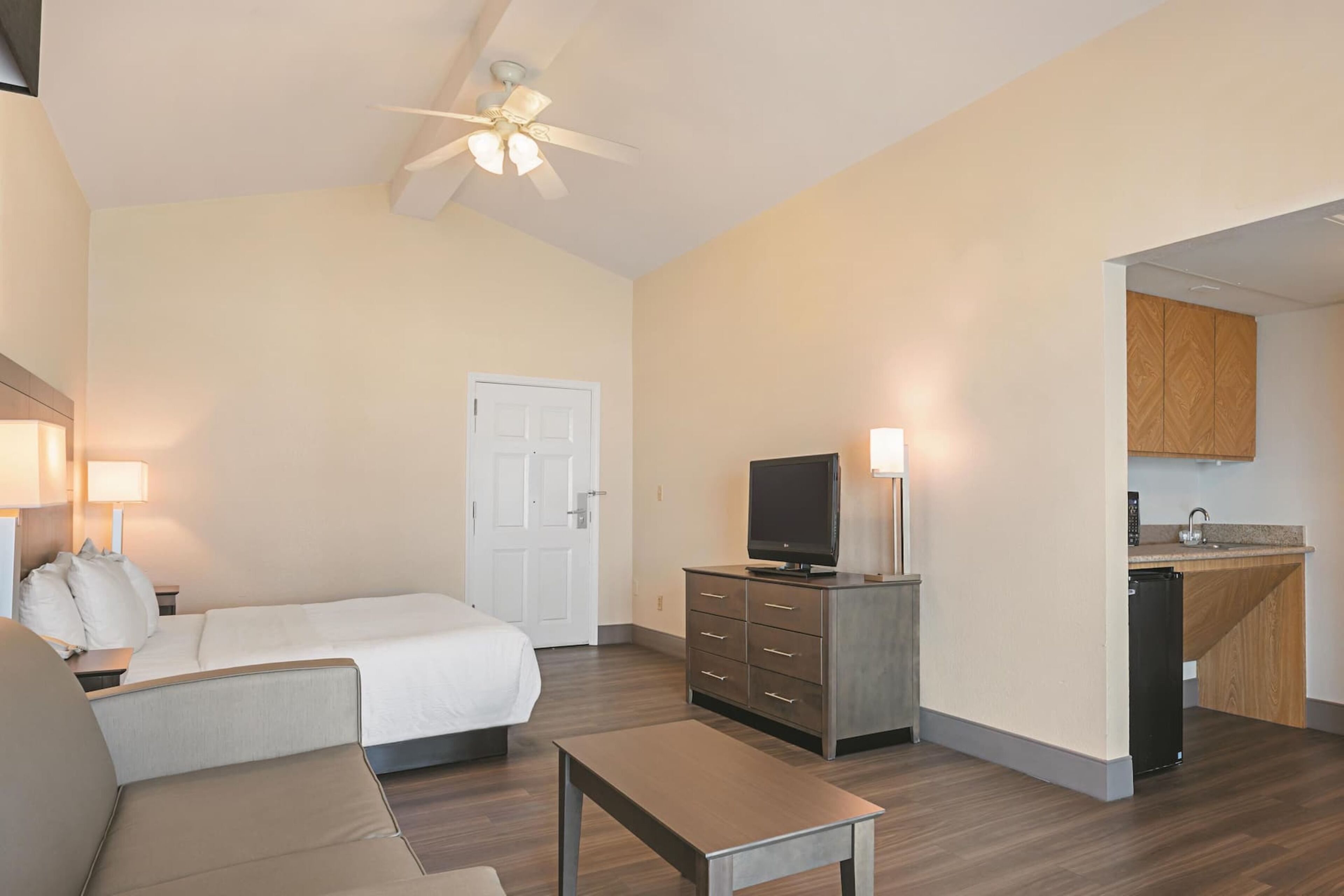 A hotel room with a bed, a desk, a television, a kitchenette, and a ceiling fan.