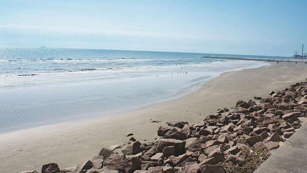 A beach with a rocky shoreline and a pier in the distance. - Wingate by Wyndham Galveston Beachfront at the Seawall (Galveston)