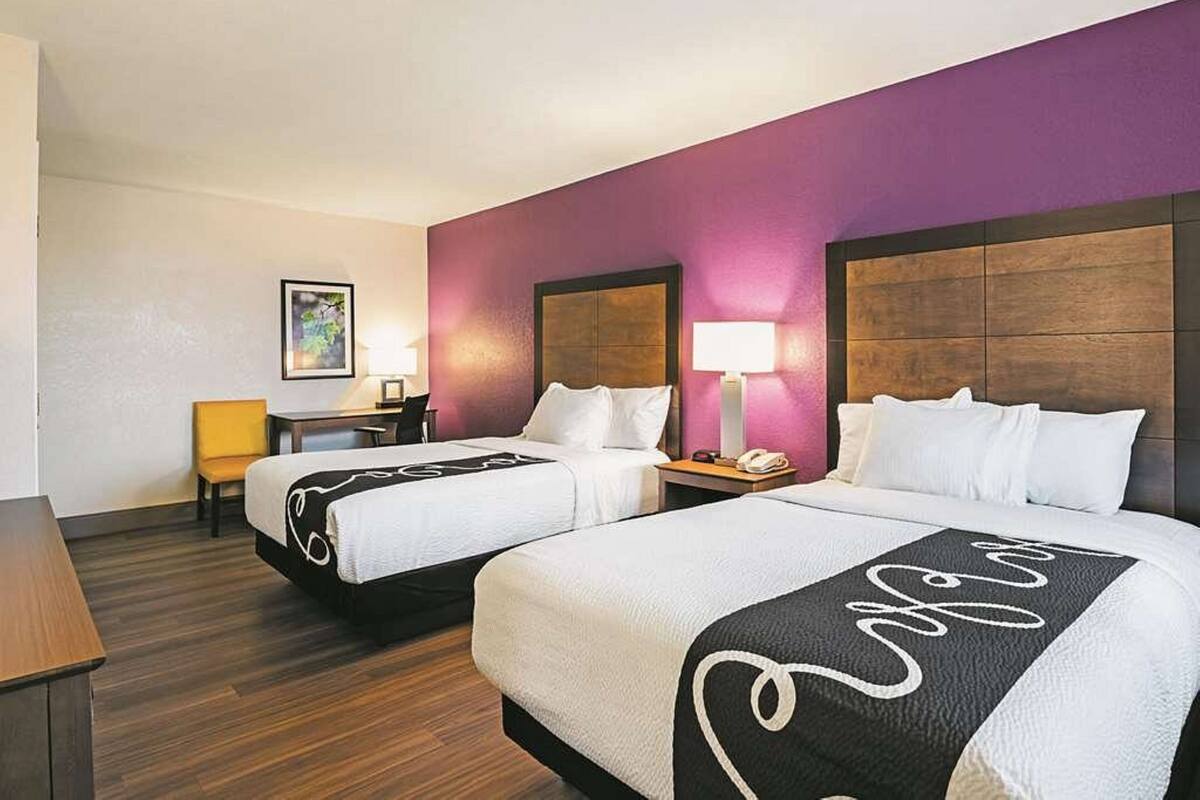 A hotel room with two beds, a desk, a chair, and a nightstand with a lamp.