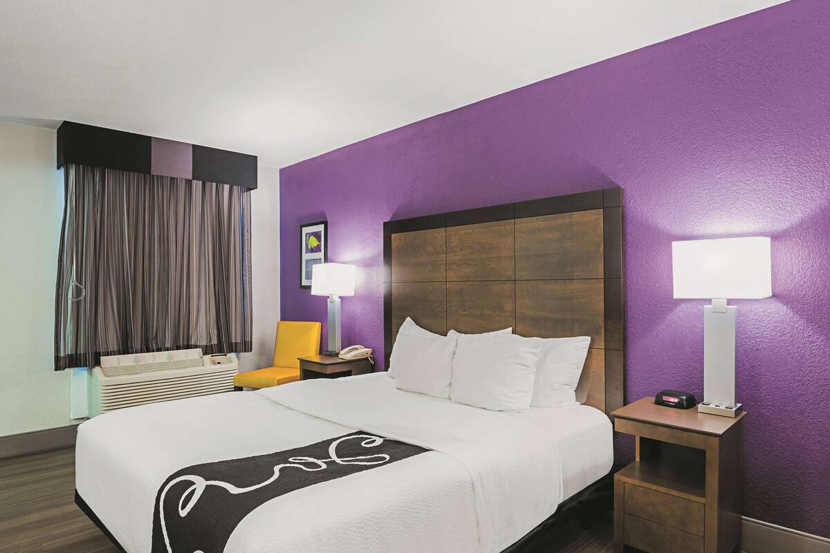 A hotel room with a large bed, a nightstand with a lamp, a chair, and a window with curtains.
