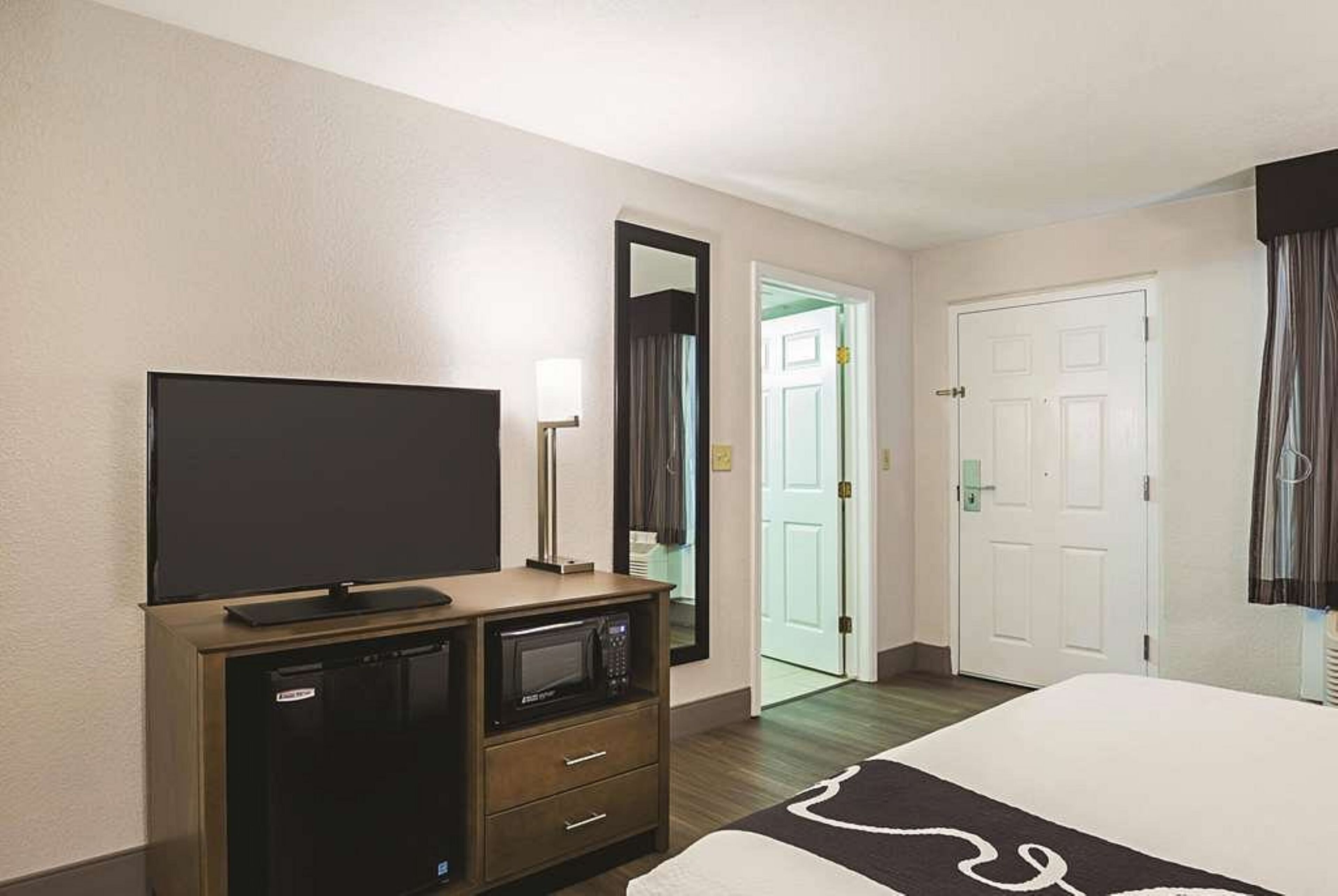 A hotel room with a flat-screen TV on a wooden dresser, a microwave, a bed with a black and white patterned bedspread, and a door leading to a bathroom.