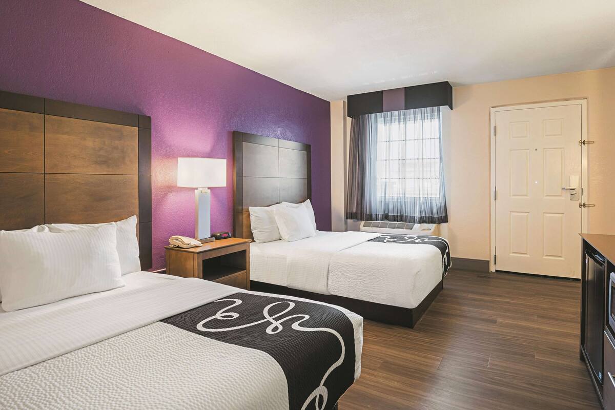 A hotel room with two beds, a nightstand with a lamp, a window with curtains, and a door.