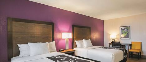 A hotel room with two beds, a desk, a chair, and a nightstand with a lamp.