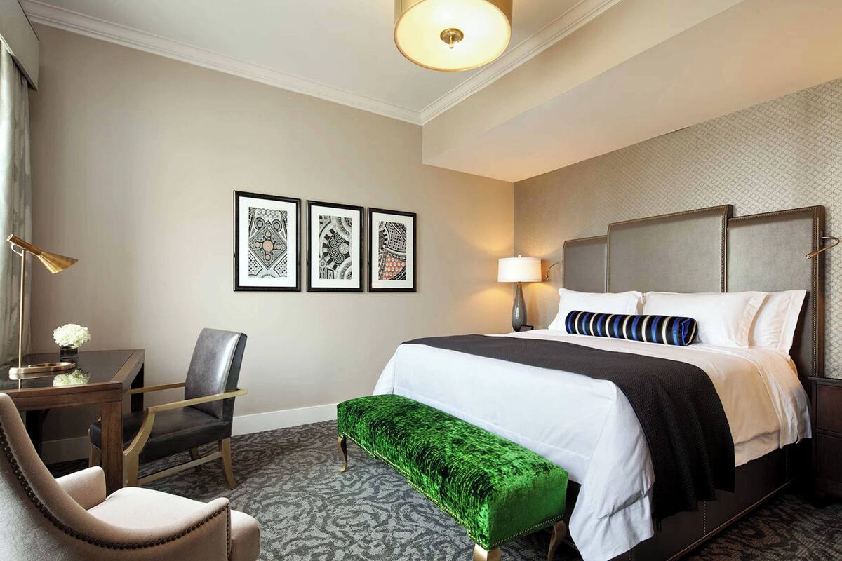 A hotel room with a bed, a desk, a chair, a bench, and framed artwork on the wall.