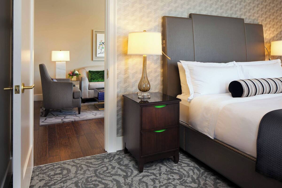 A hotel room with a bed, a nightstand with a lamp, a chair, and a small table with a vase.