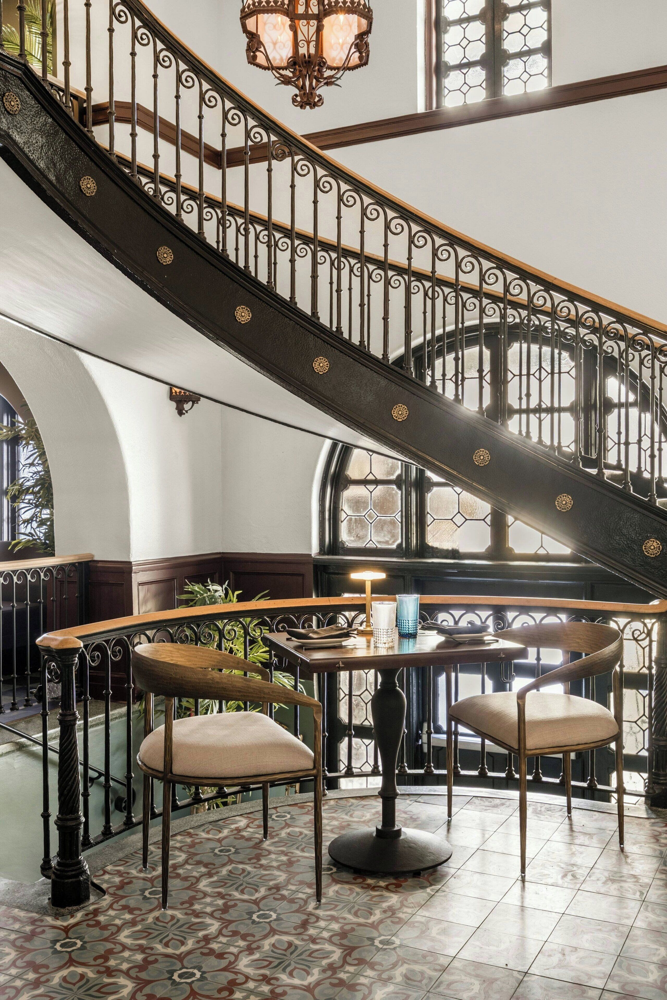 A grand staircase with ornate metal railings and a round table with chairs in a sunlit room with patterned flooring.