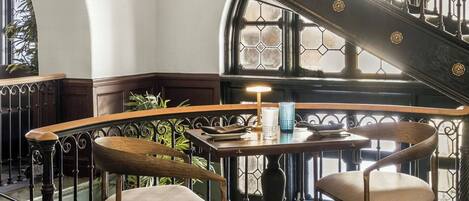 A grand staircase with ornate metal railings and a round table with chairs in a sunlit room with patterned flooring.
