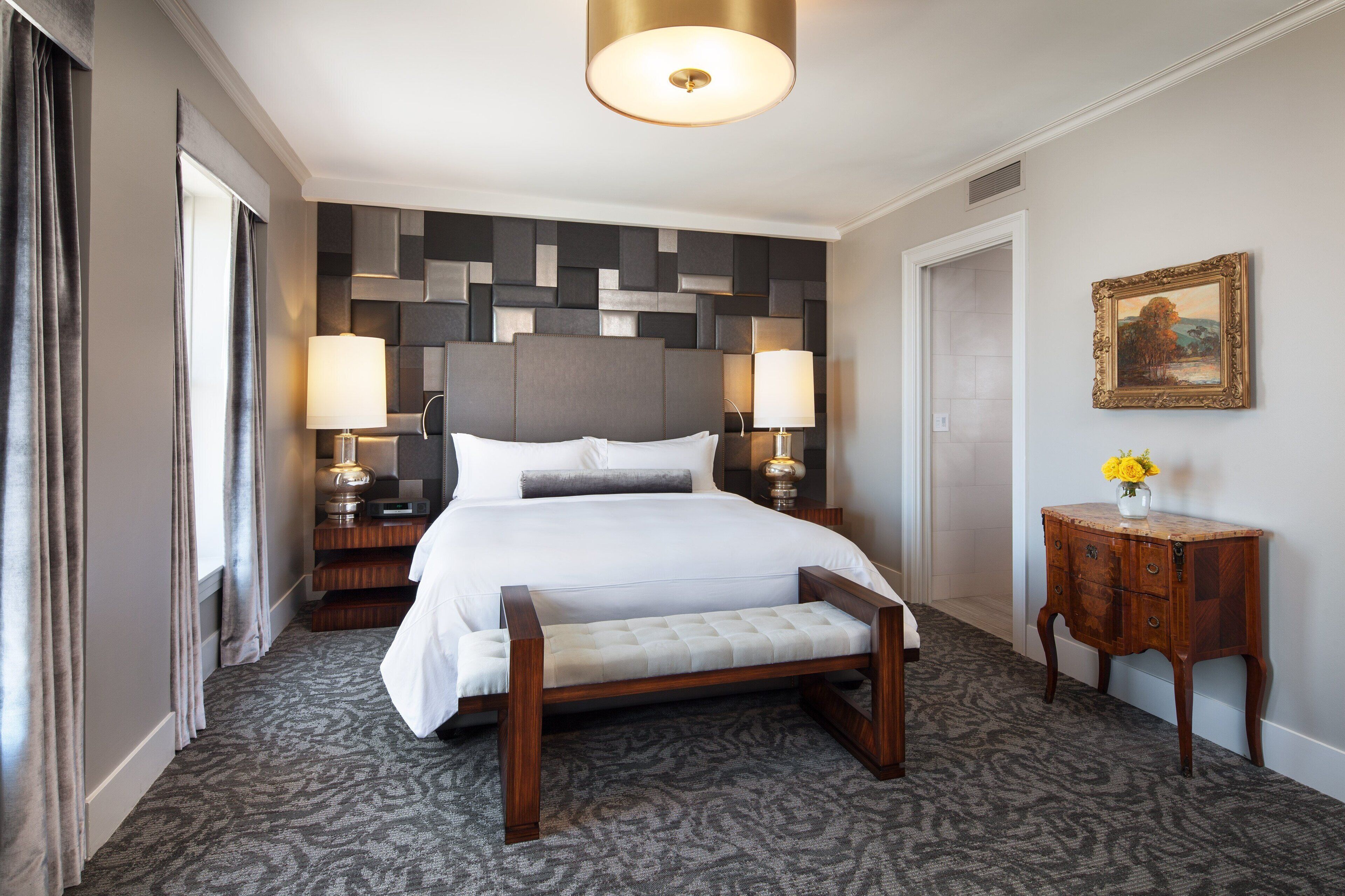 Ein Hotelzimmer mit einem großen Bett, zwei Nachttischlampen, einer hölzernen Bank, einem kleinen Tisch mit einer Vase und einem Gemälde an der Wand.