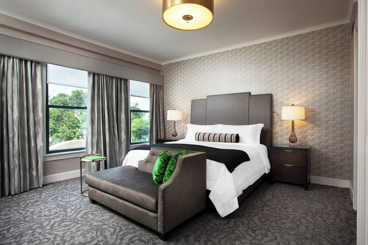 A hotel room with a large bed, two bedside lamps, a sofa, and a view of trees through the window.