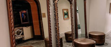 A hallway with ornate mirrors and leather ottomans, a framed painting on the wall, and a glimpse of a room with a bed and a nightstand.