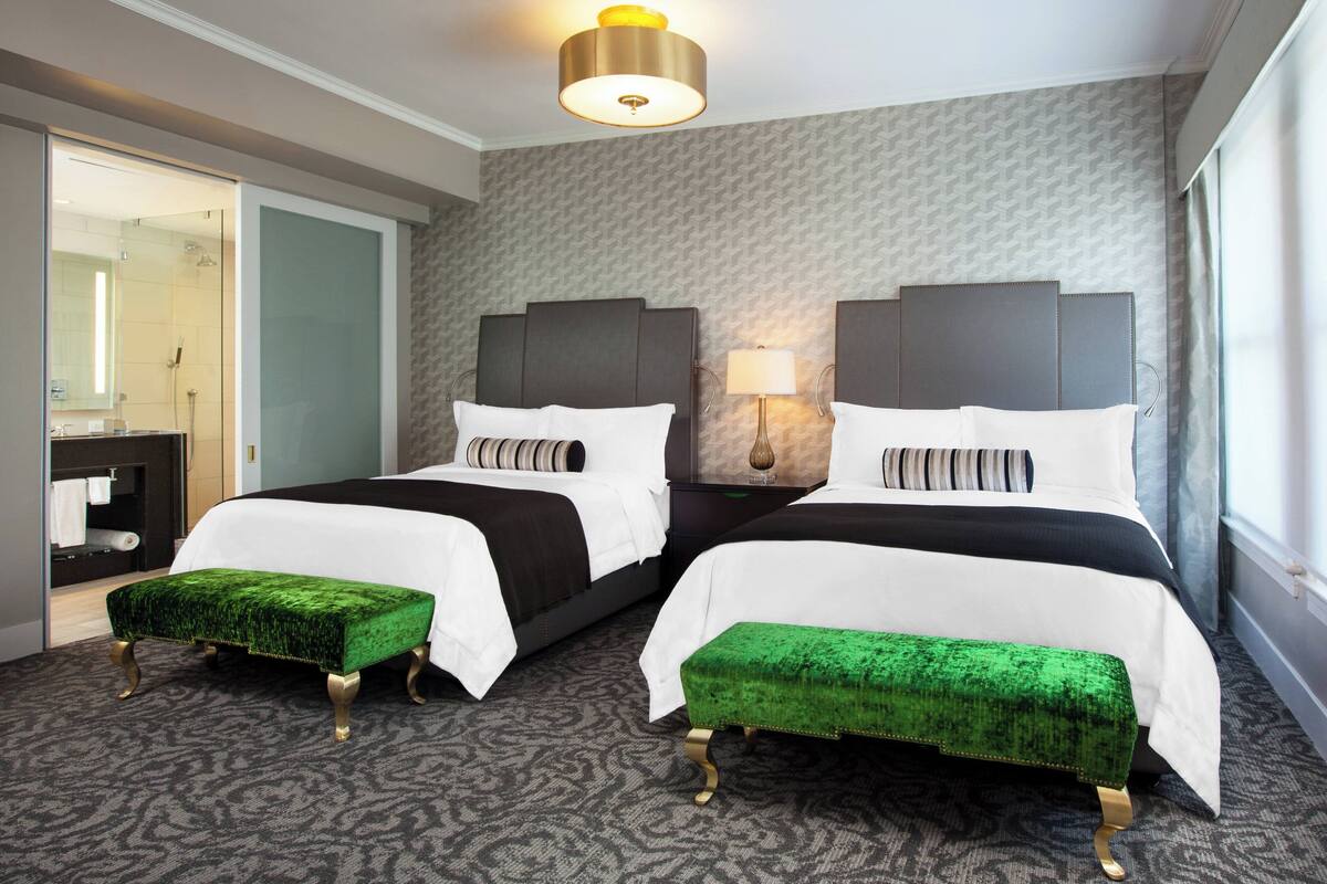 A hotel room with two beds, a green upholstered bench, a bedside table with a lamp, and a shower area visible through a glass door.