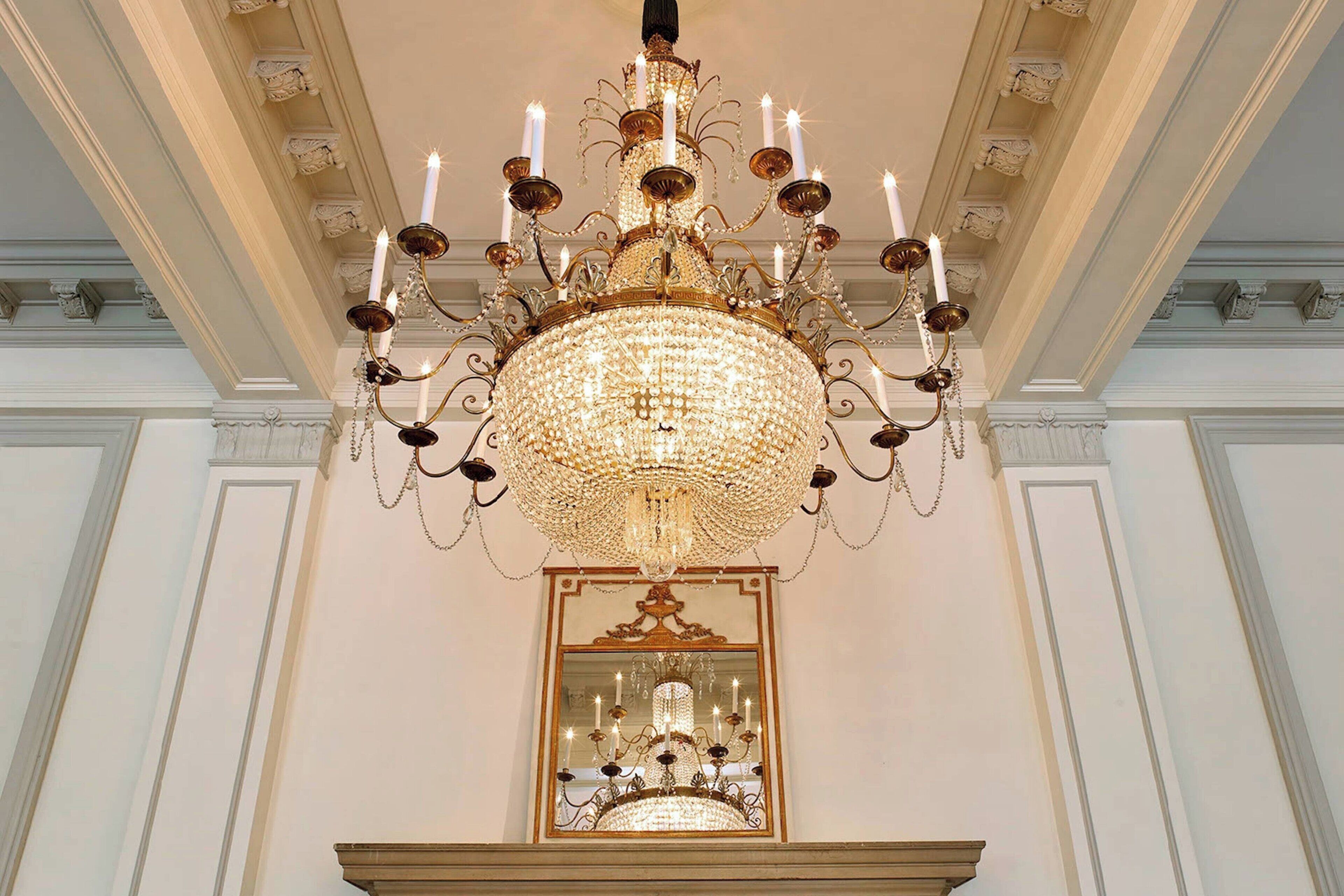 En storslagen kristallkrona med många ljus, en spegel under den och klassiska arkitektoniska detaljer i taket.