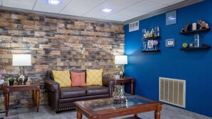 A living room with a stone wall, a blue accent wall, a brown leather sofa with yellow and red cushions, a glass coffee table, and two wooden side tables with lamps. - Days Inn & Suites by Wyndham Commerce (Commerce)