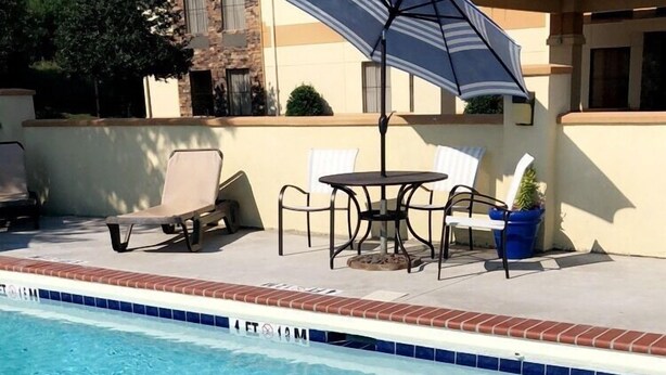 A clear swimming pool with a tiled edge, a sun umbrella, and outdoor seating with chairs and a table. - Days Inn & Suites by Wyndham Commerce (Commerce)