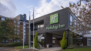 A Holiday Inn hotel with a modern design, featuring a prominent logo, a staircase leading to the entrance, and a parking area with marked spaces.
