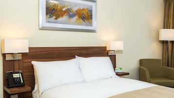 A hotel room with a wooden headboard, white bedding, a bedside phone, a framed abstract painting, and a green armchair.