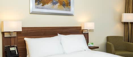 A hotel room with a wooden headboard, white bedding, a bedside phone, a framed abstract painting, and a green armchair.