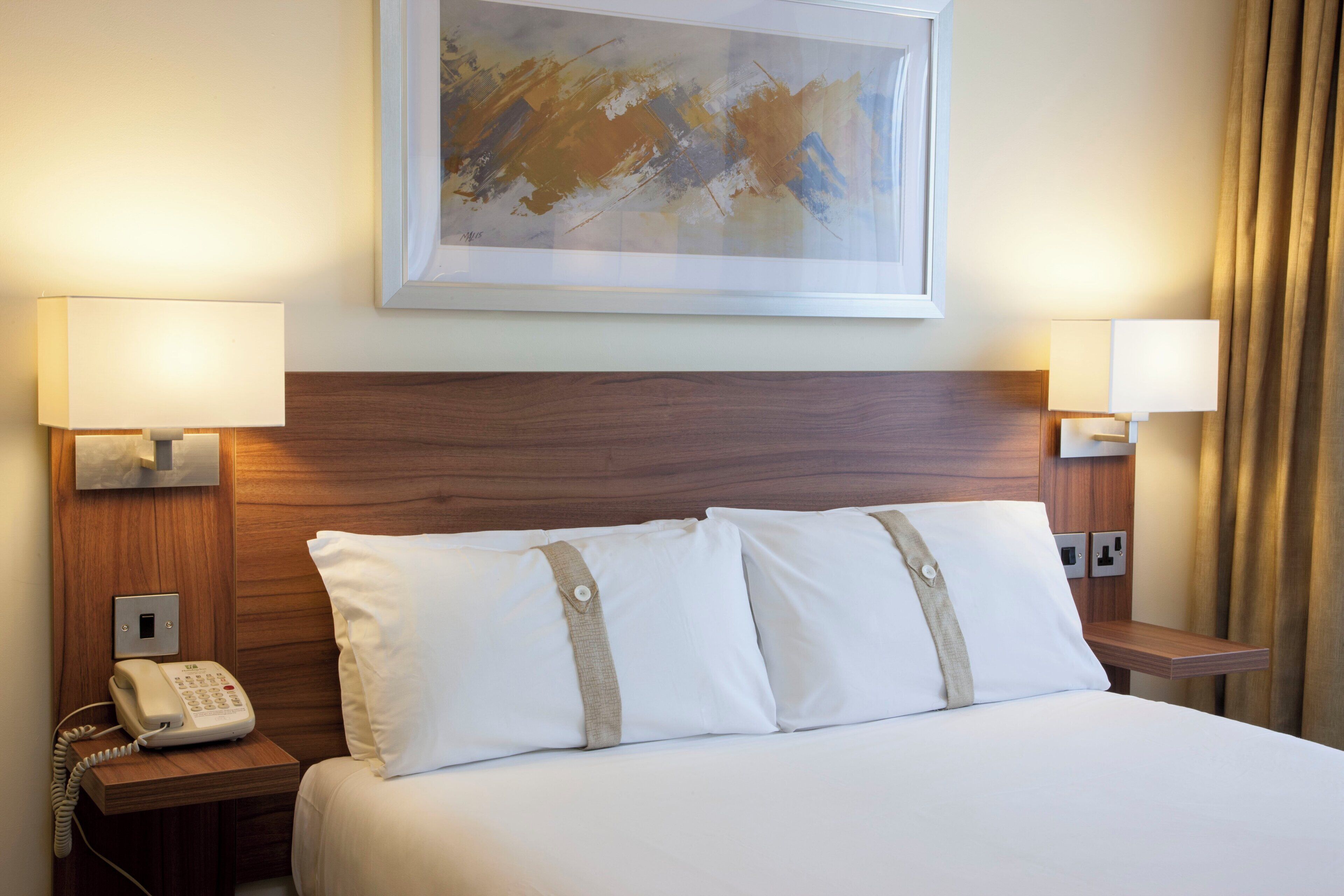 A hotel room with a bed, two bedside lamps, a telephone, and a painting on the wall.