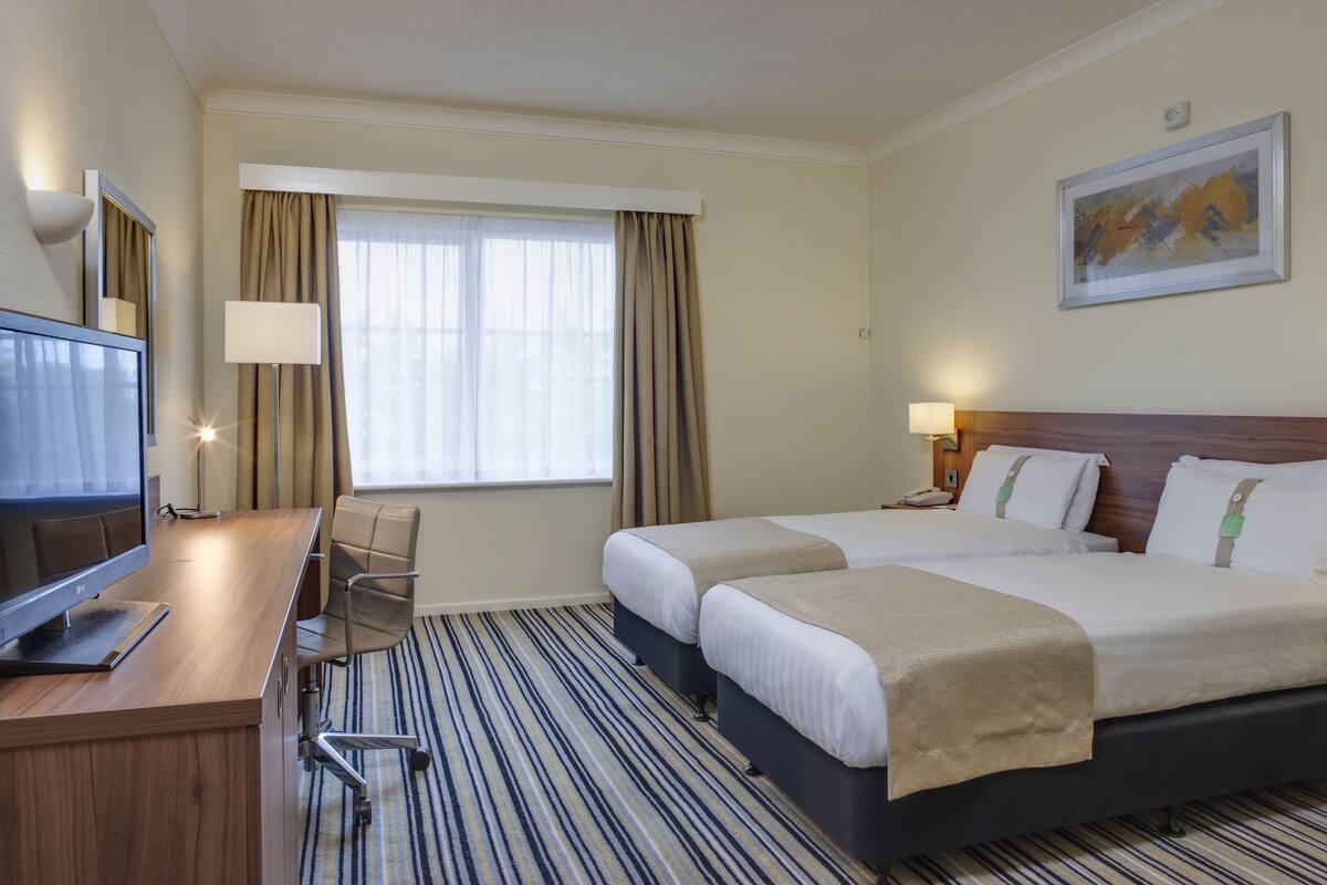 Ett hotellrum med ett skrivbord, två sängar, en tv och ett fönster med gardiner.