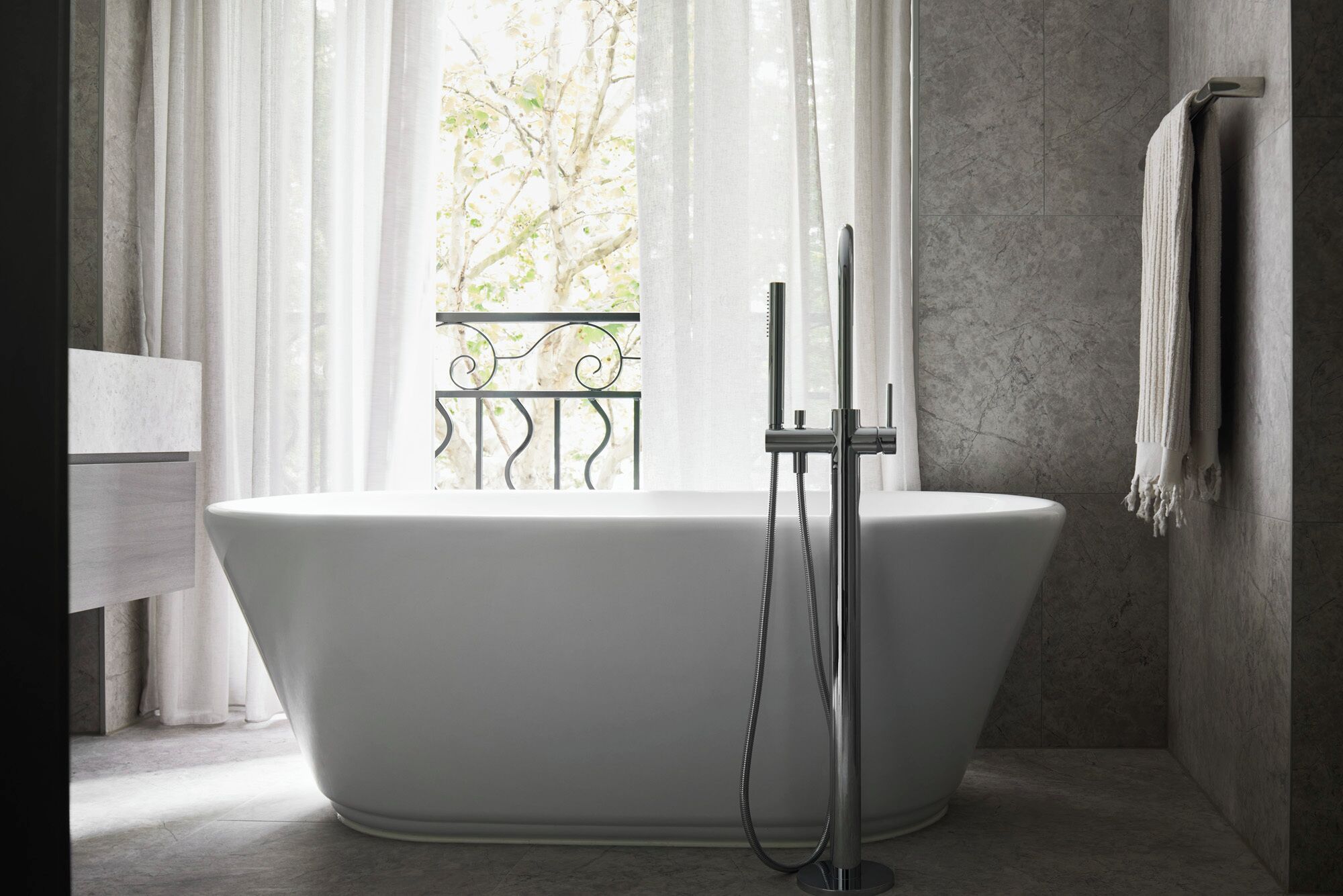 A modern bathroom with a freestanding bathtub, a towel rack, and a view through a window with a balcony.