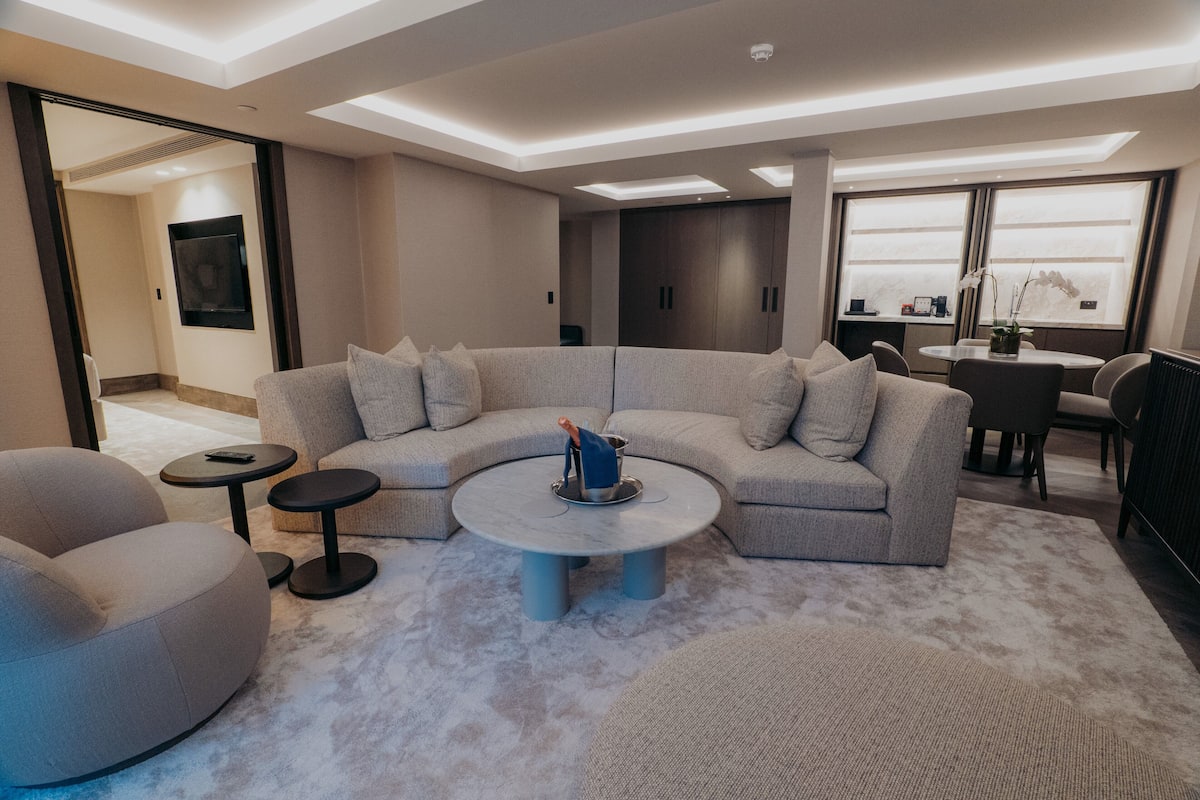 A modern living room with a sectional sofa, a round coffee table, and a small side table. There is a dining area with a table and chairs visible in the background.