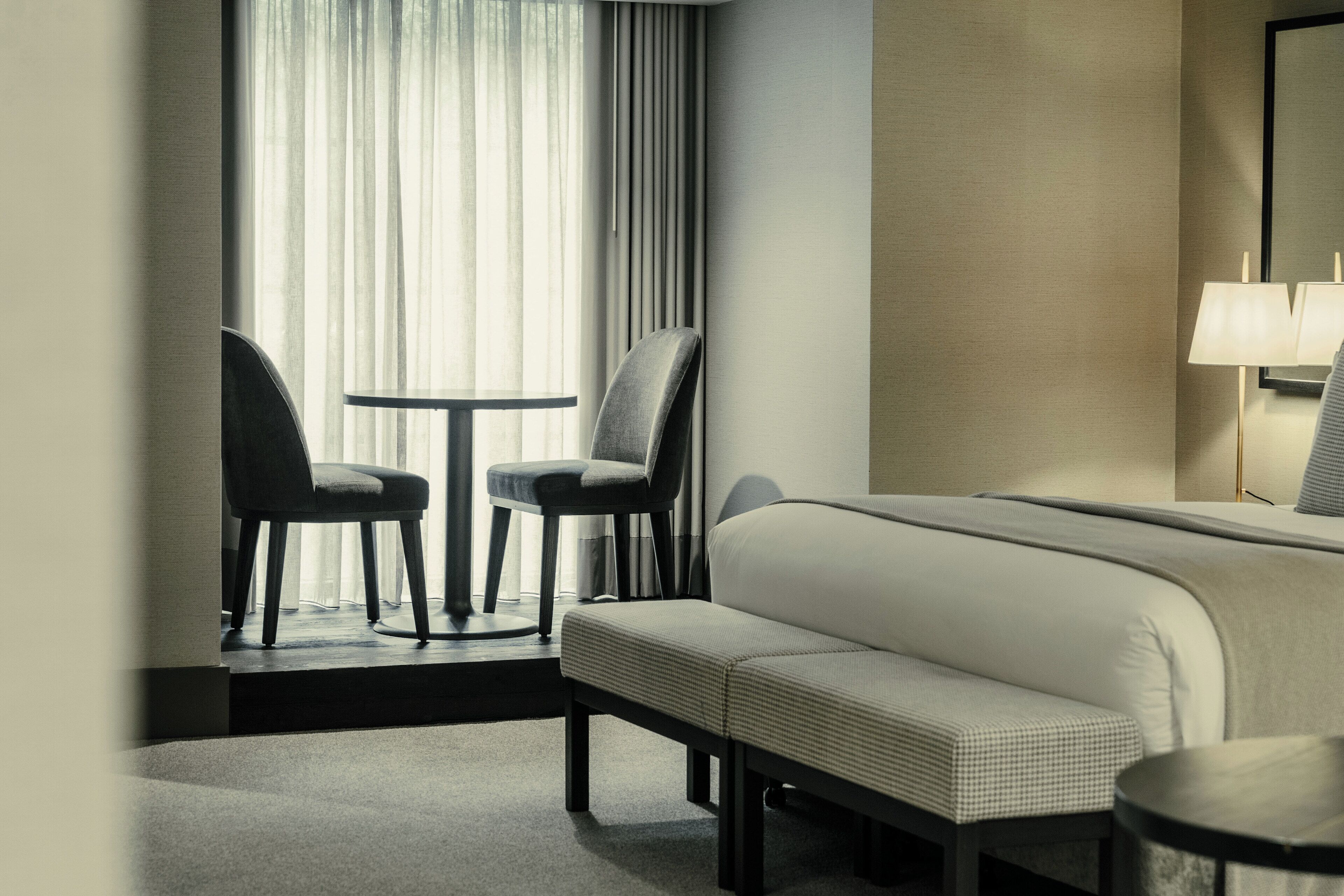 A hotel room with a bed, two chairs, a small table, a bench, and a lamp.