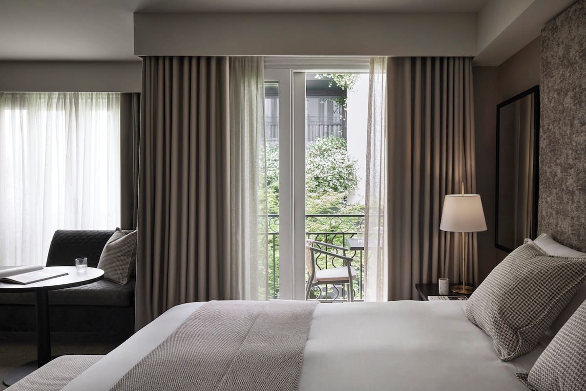 A hotel room with a bed, a desk, a chair, a lamp, and a view of a balcony with greenery.