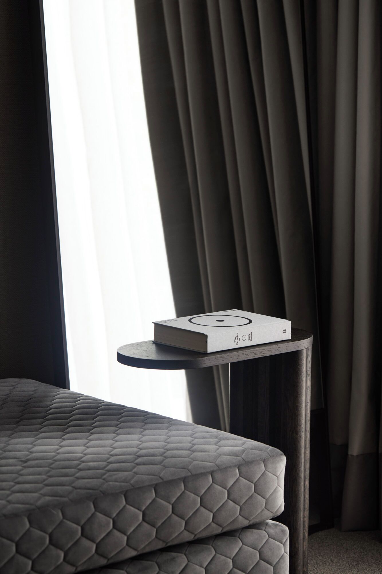 A neatly made bed with a geometric patterned cover, a small bedside table with a book, and a window with curtains.
