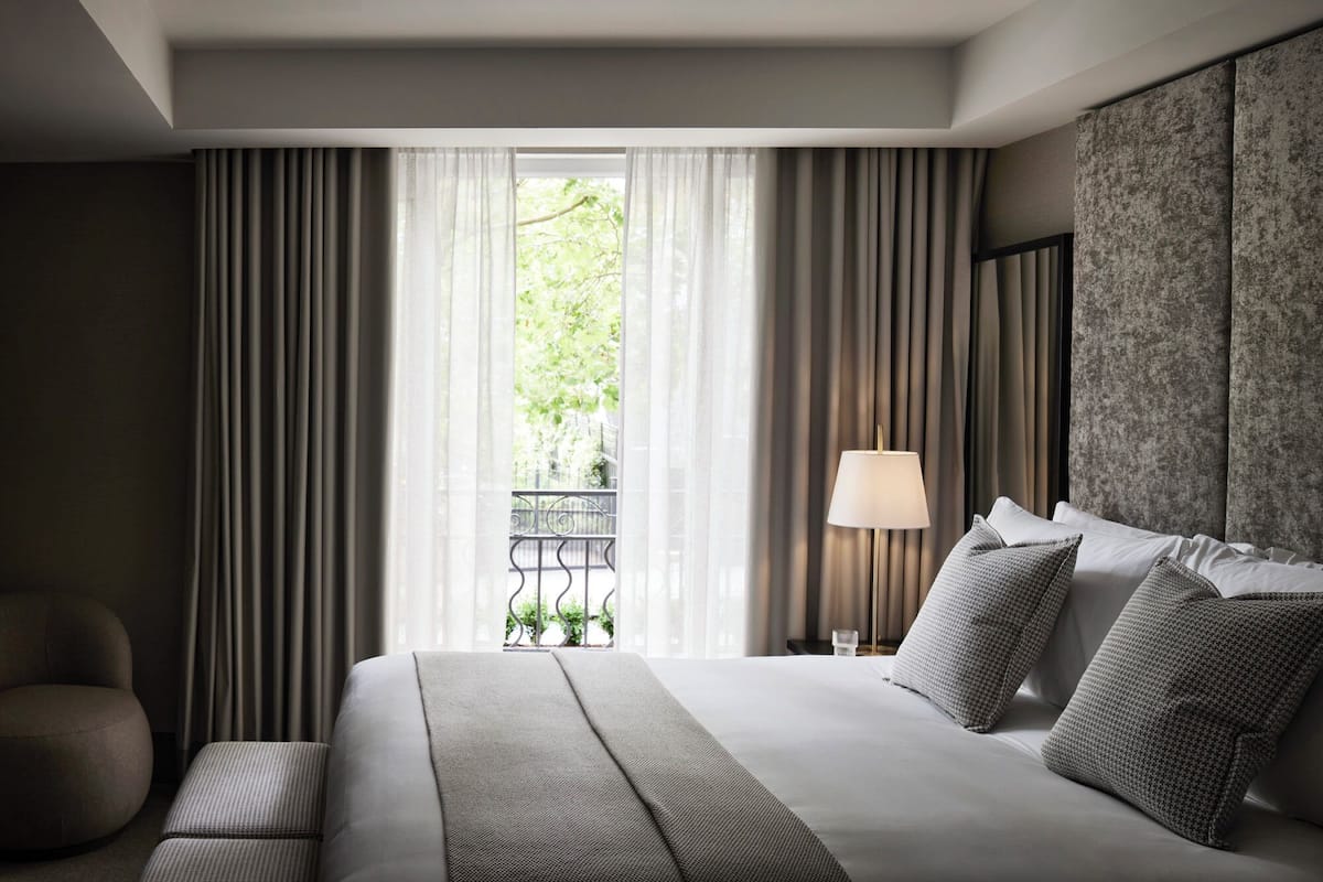 A hotel room with a large bed, a bedside table with a lamp, and a view of greenery through sheer curtains.