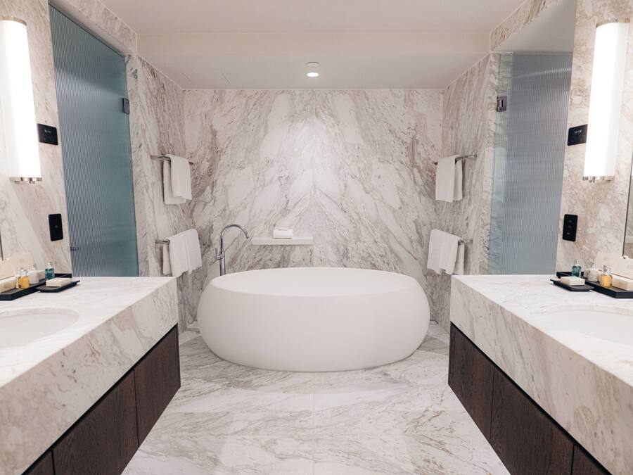 Un baño moderno con una bañera ovalada grande, dos lavamanos con armarios de madera oscura y paredes de mármol.
