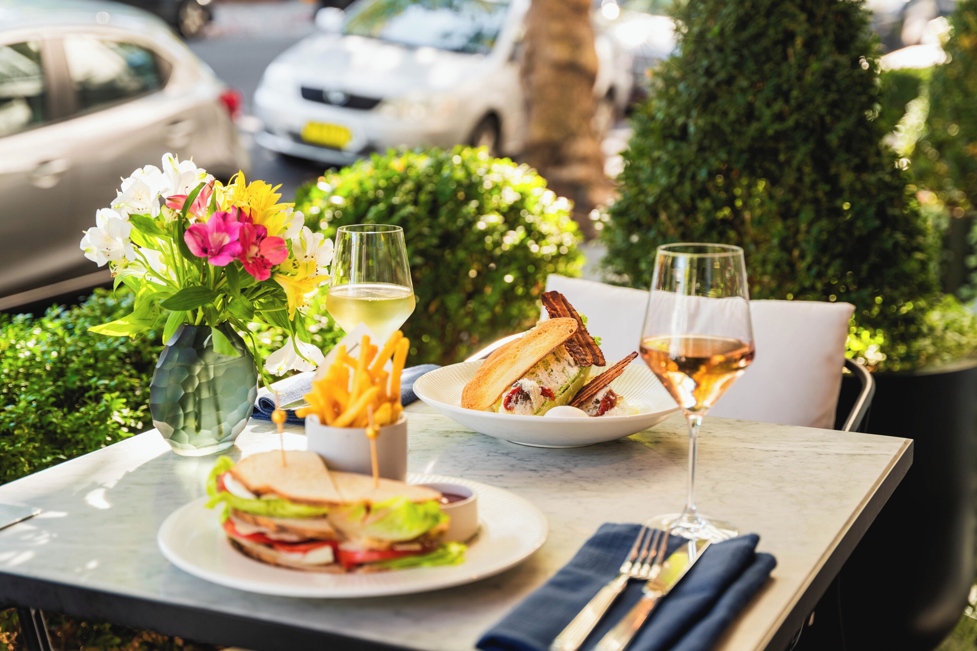 A table set with a sandwich, fries, a glass of white wine, and a vase of flowers.