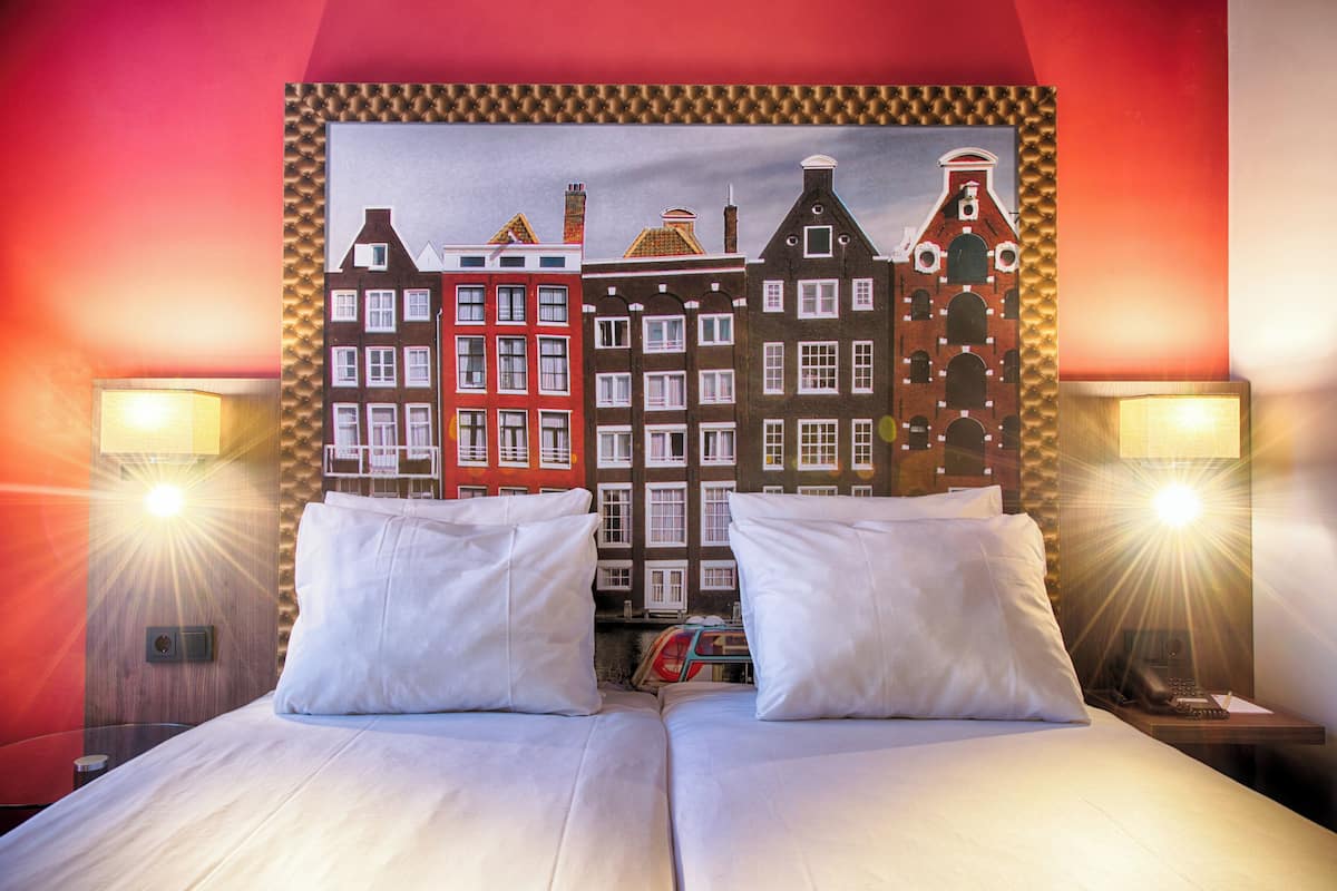 A hotel room with a bed, two bedside tables, and a wall painting of European buildings.