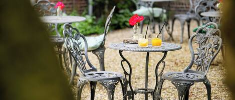 Mobiliario de patio exterior con sillas y mesas metálicas, una mesa pequeña con un libro y un vaso de jugo de naranja, y una rosa en un florero.