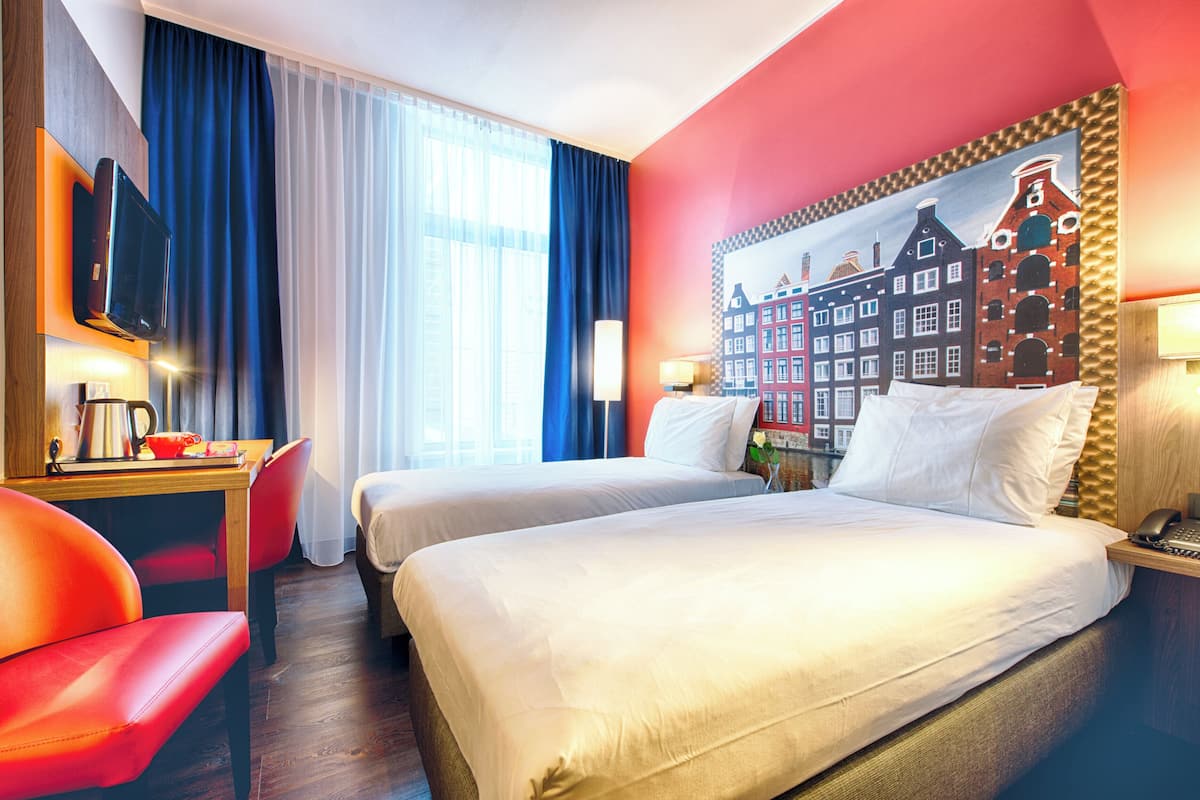 A hotel room with two beds, a desk with a chair, a TV, and a painting of a cityscape on the wall.