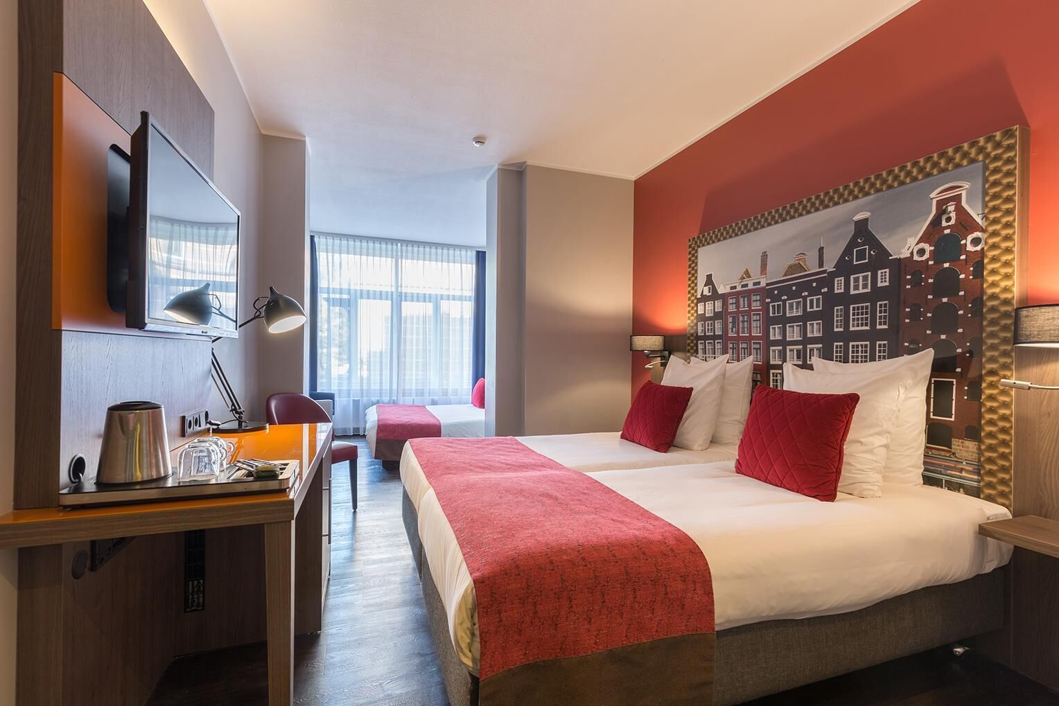 Quarto de hotel com cama, escrivaninha com chaleira, televisão e pintura de uma paisagem urbana.
