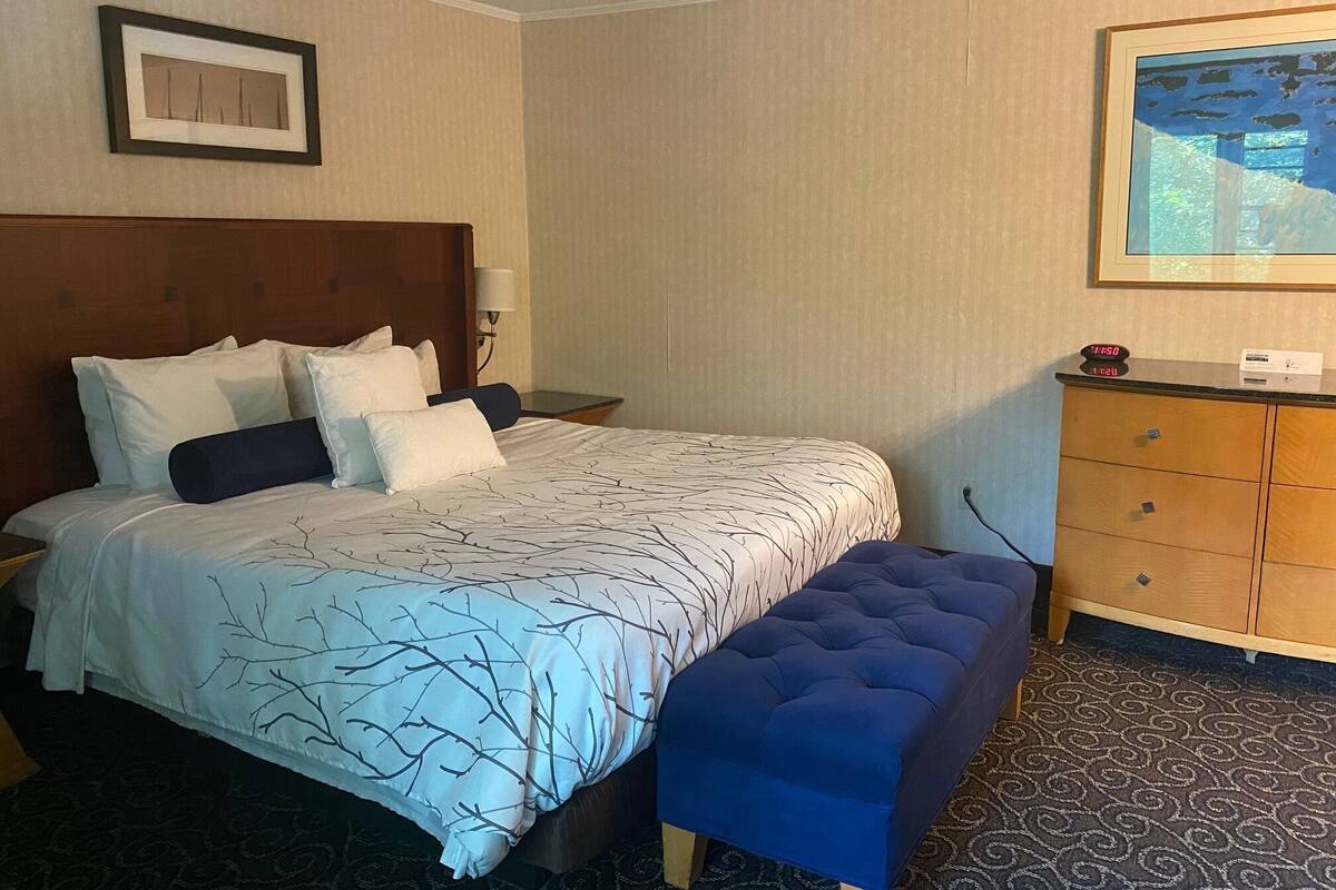 A neatly made bed with a patterned bedspread, a wooden headboard, a bedside table with a lamp, a wooden dresser with a mirror, and a framed picture on the wall.