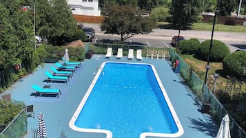 A rectangular swimming pool with a blue border, surrounded by lounge chairs and umbrellas, with a fence and greenery in the background.