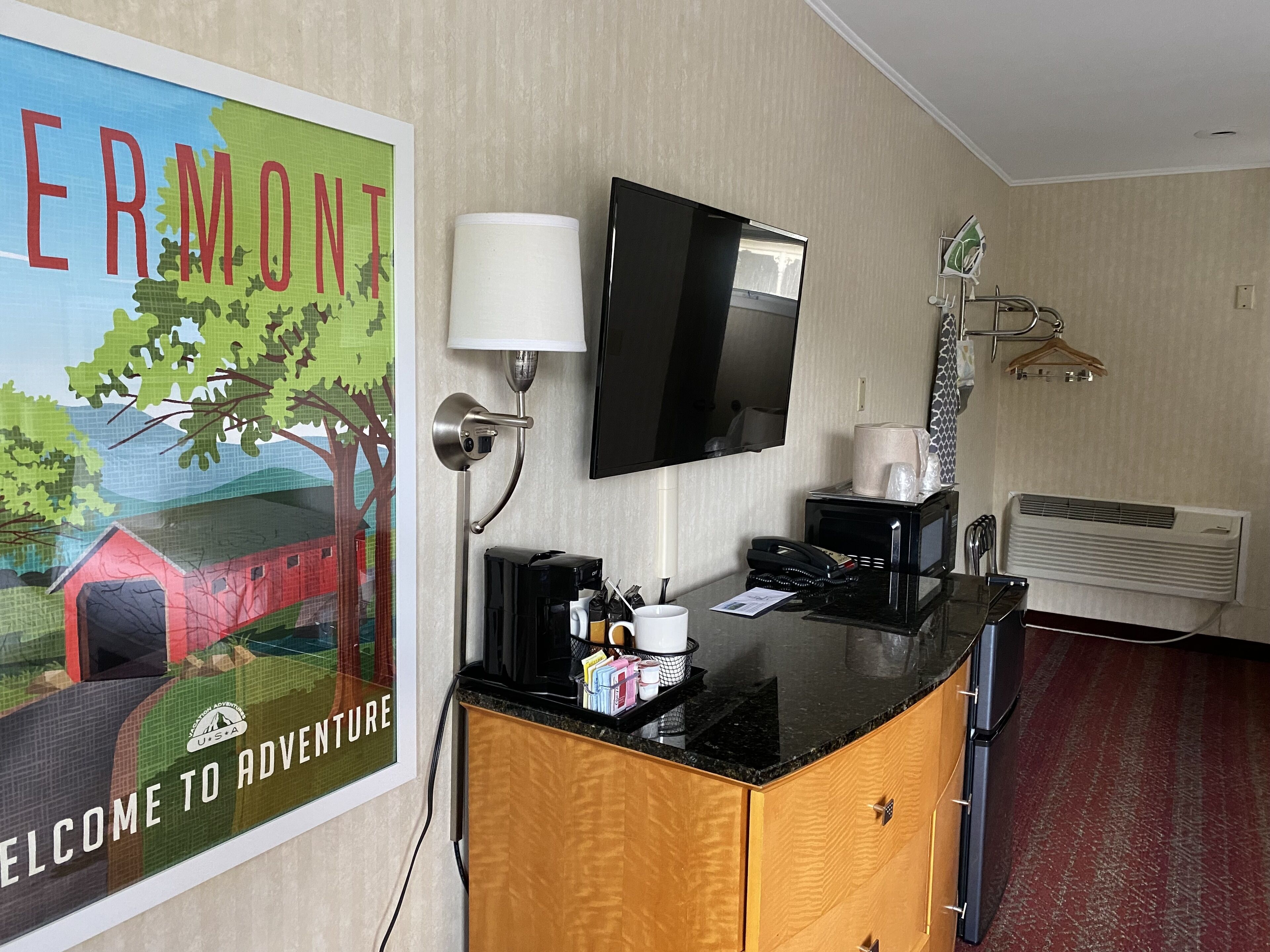 Hotellihuone, jossa on sänky, työpöytä tietokoneella, televisio, lamppu, kahvinkeitin ja juliste seinällä.