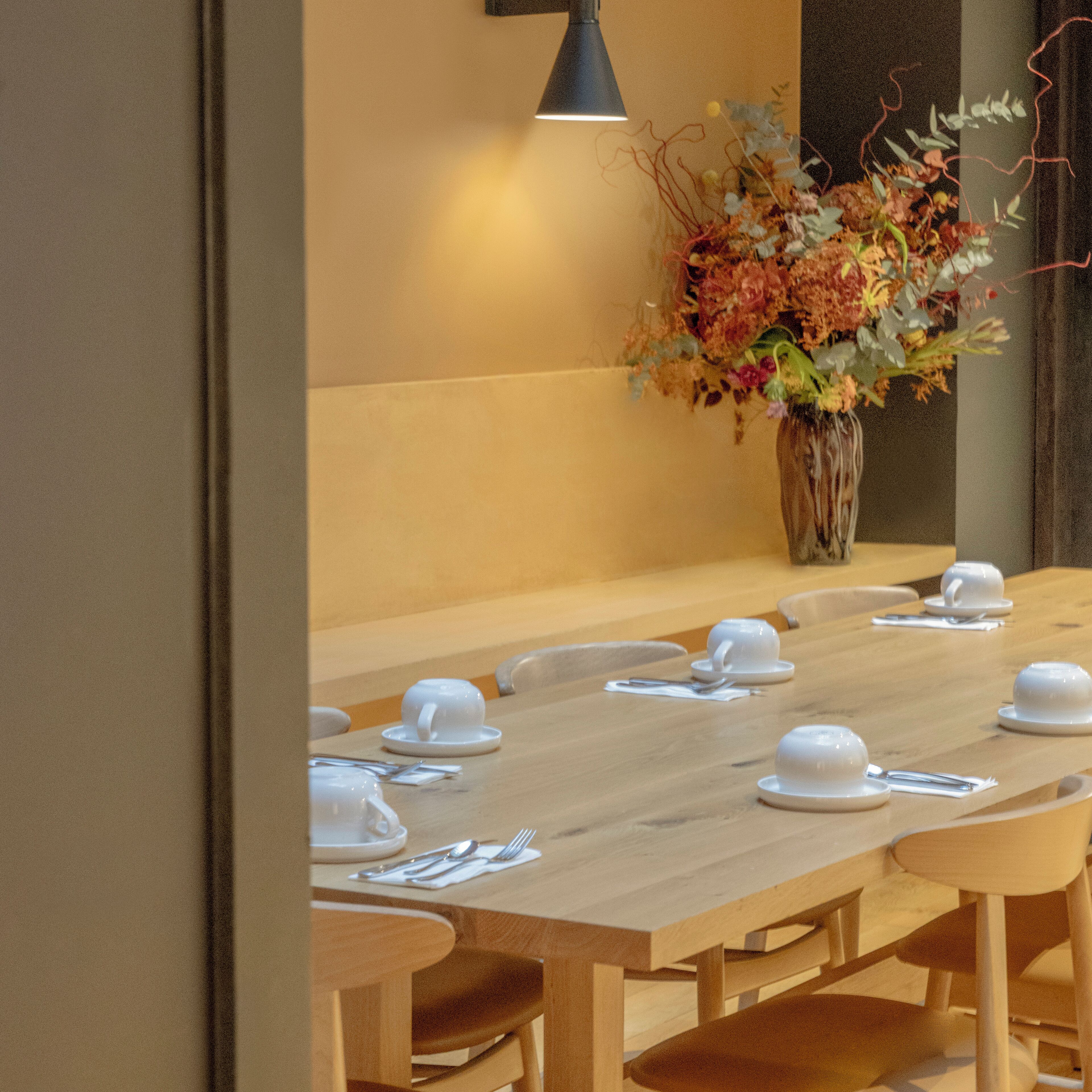 A dining area with a long wooden table set for a meal, a vase of flowers, and a wall-mounted lamp.
