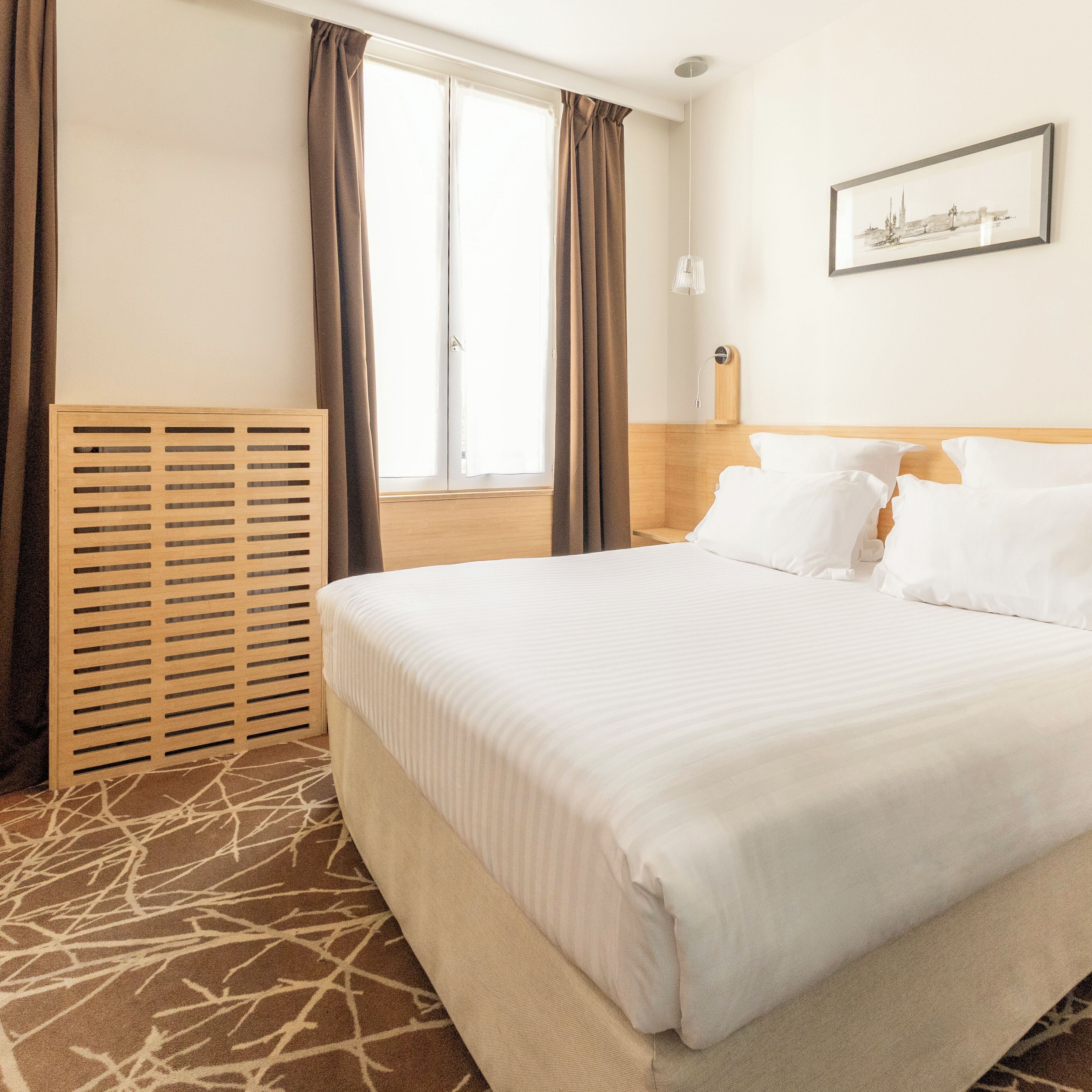 A hotel room with a large bed, a wooden headboard, a window with curtains, a wall-mounted lamp, and a framed picture.