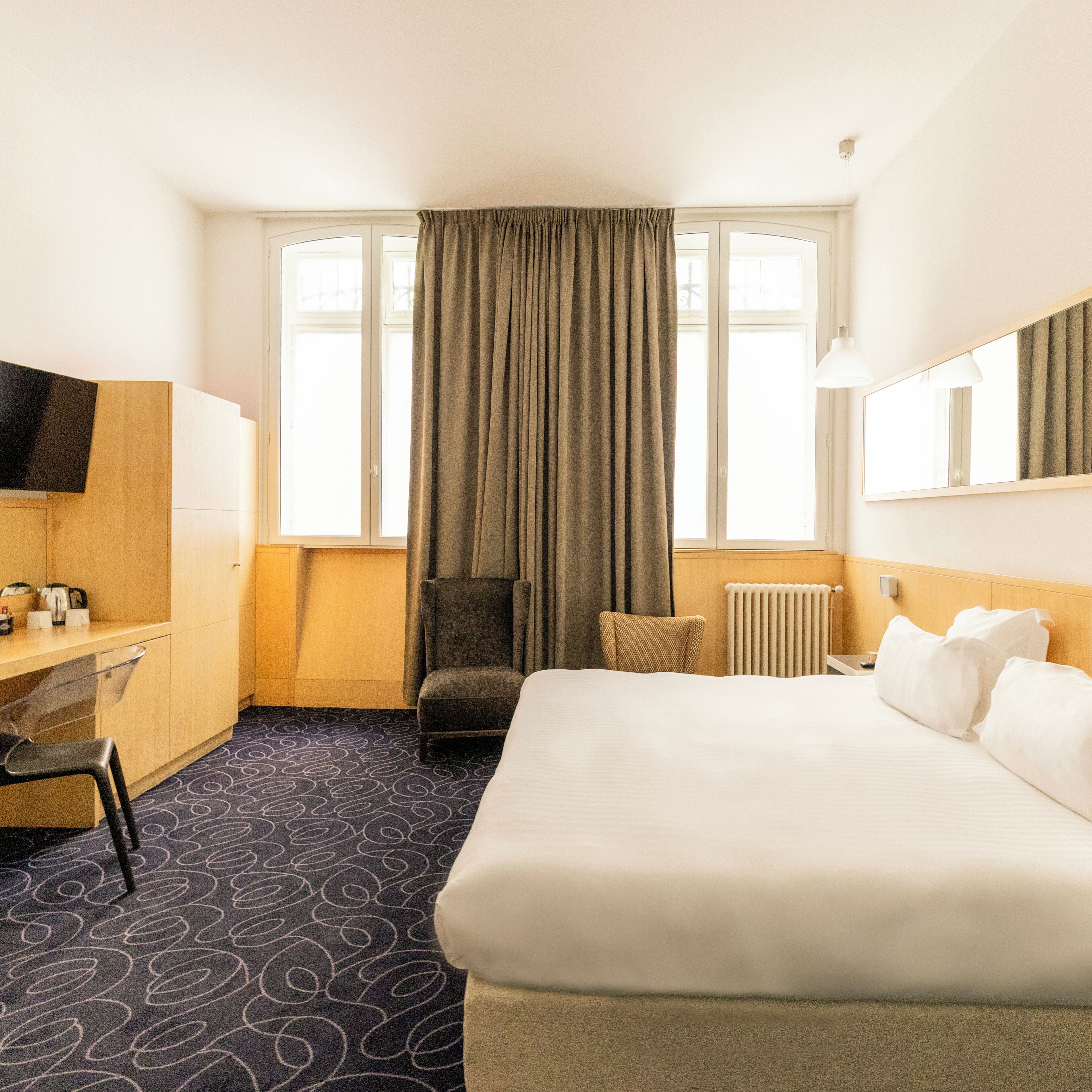 Quarto de hotel com cama, escrivaninha, cadeira, televisão e janela com cortinas.