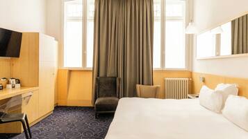 Quarto de hotel com cama, escrivaninha, cadeira, televisão e janela com cortinas.