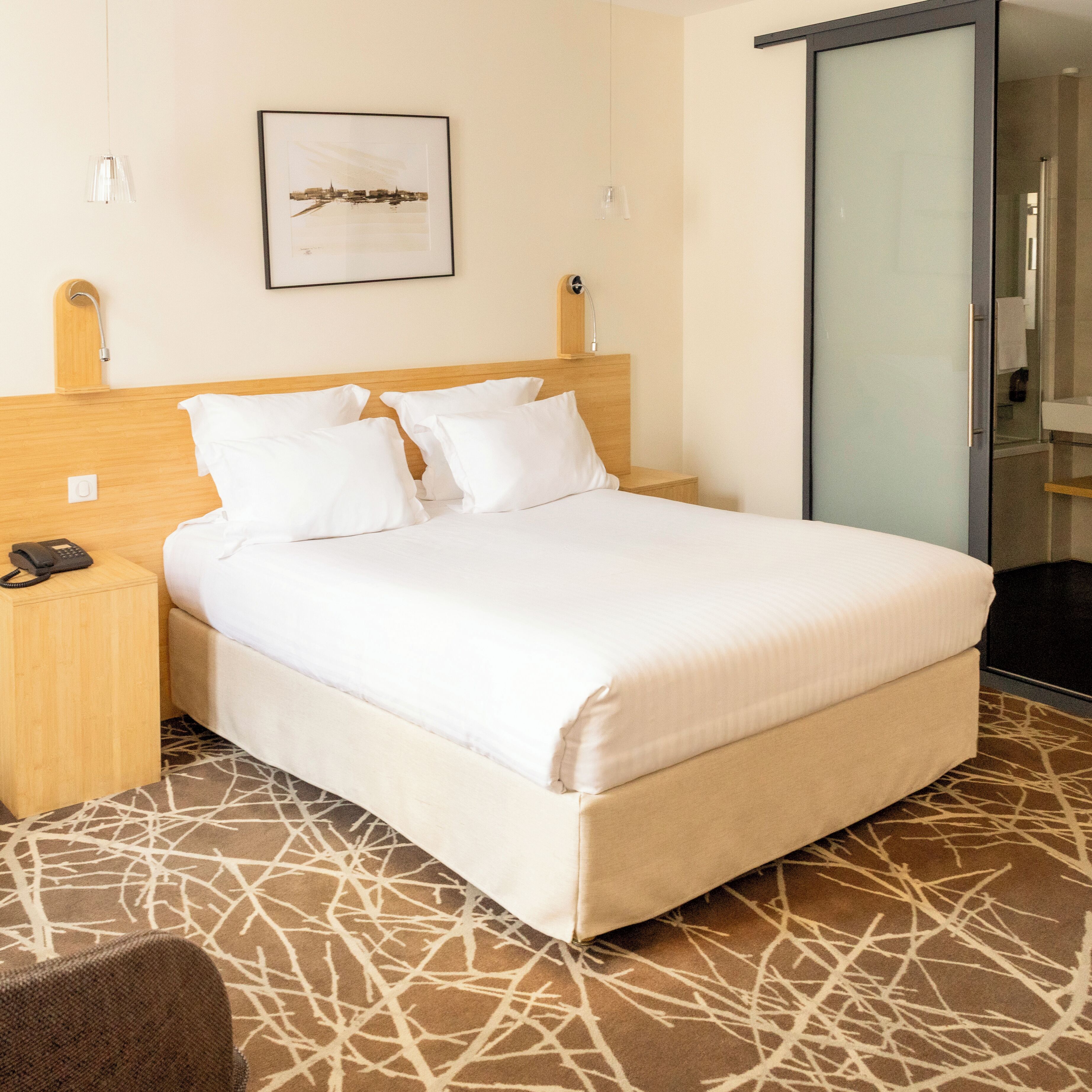 A hotel room with a bed, a bedside table with a phone, a framed picture on the wall, and a bathroom door.