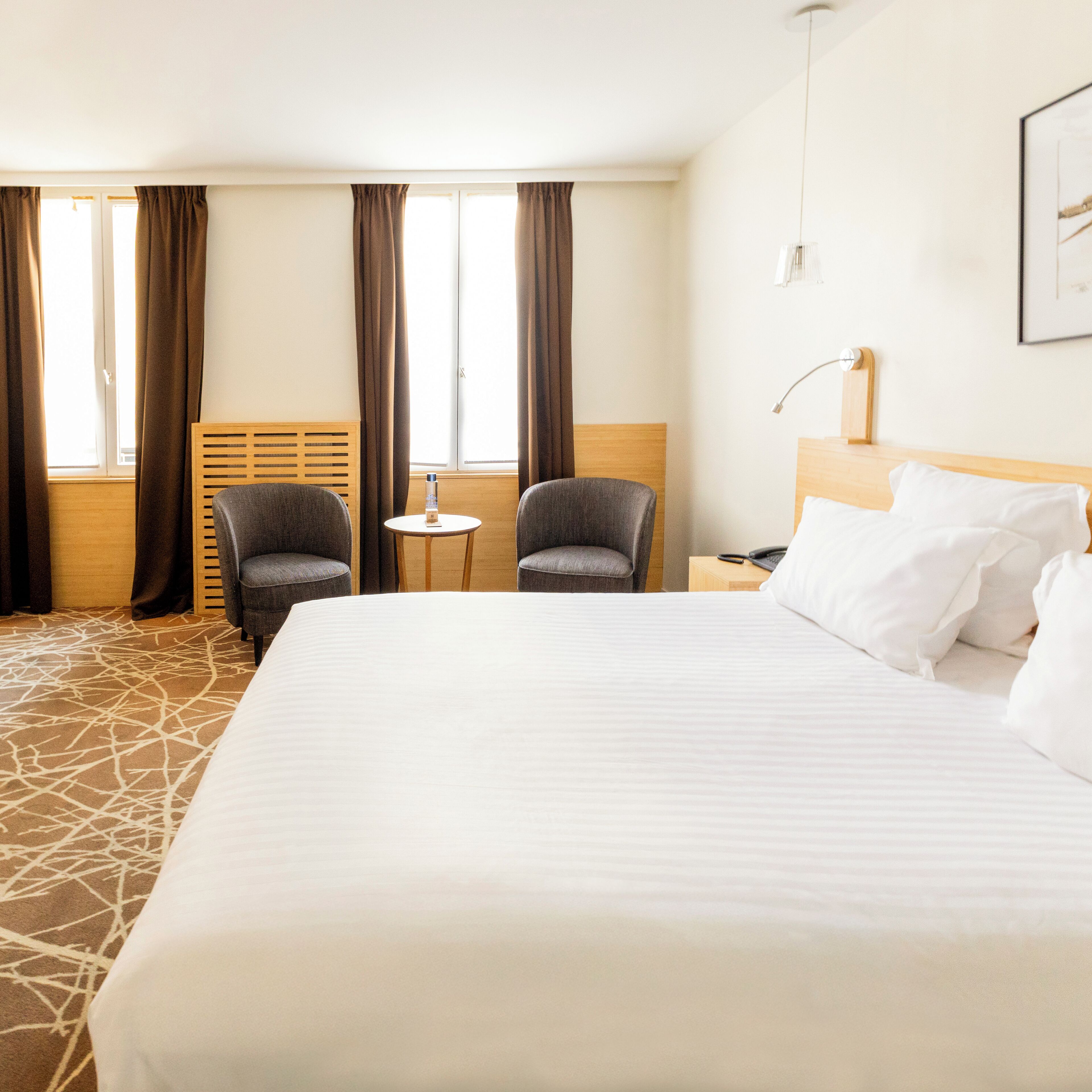 Habitación de hotel con una cama grande, dos sillones, una mesita y un ventanal con cortinas.