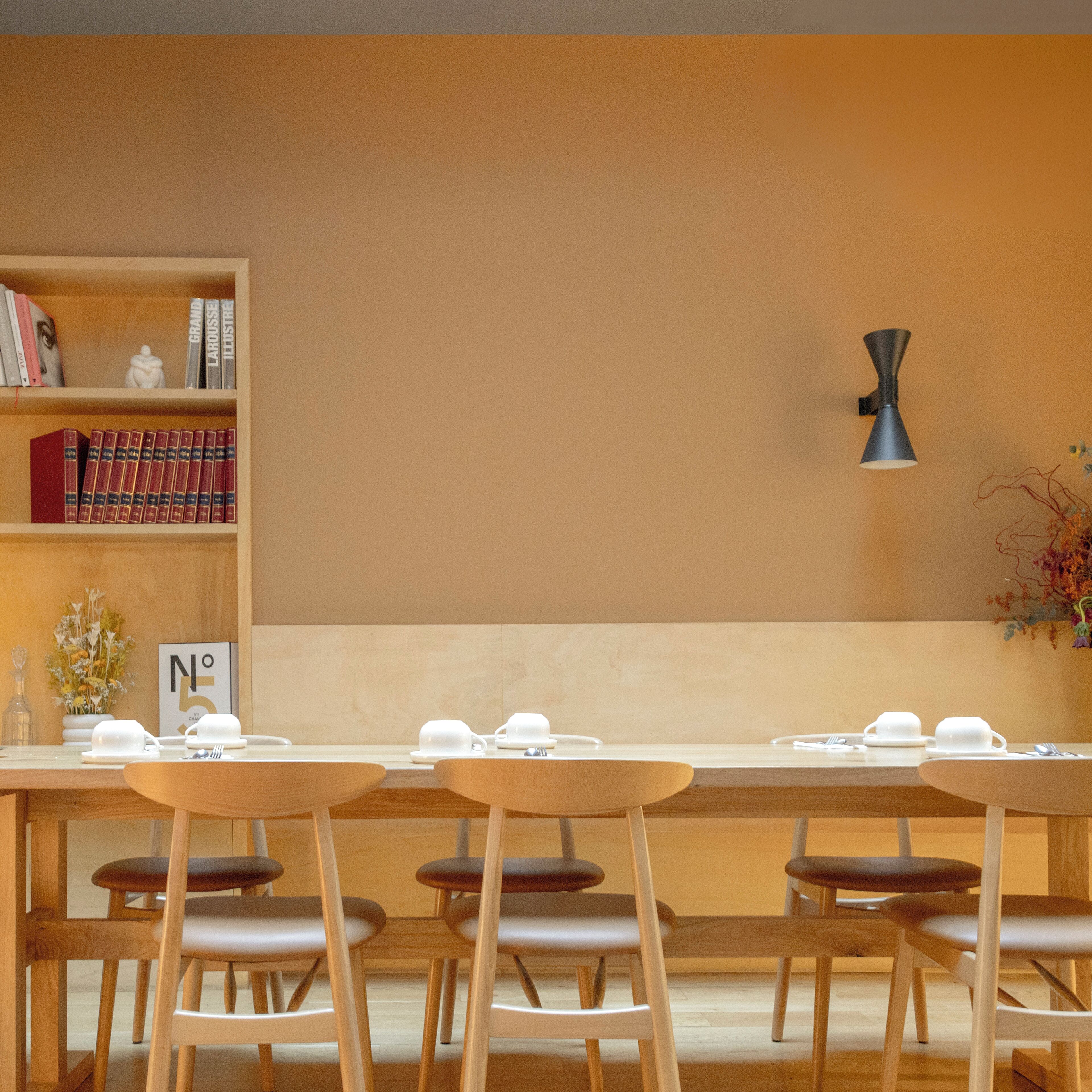 Un comedor con una mesa larga dispuesta para seis personas, sillas de madera, una estantería con libros, una lámpara fijada a la pared y un letrero que dice 